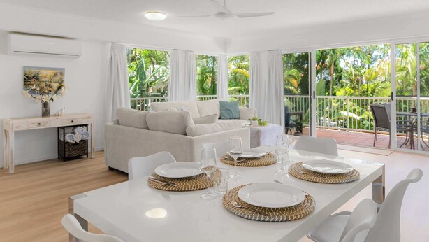 A bright, airy room with a white table set for dining, a sofa, and a view of lush greenery outside. - Noosa Hill Getaway - Walk to Beach + Shared Pool (Noosa Region)