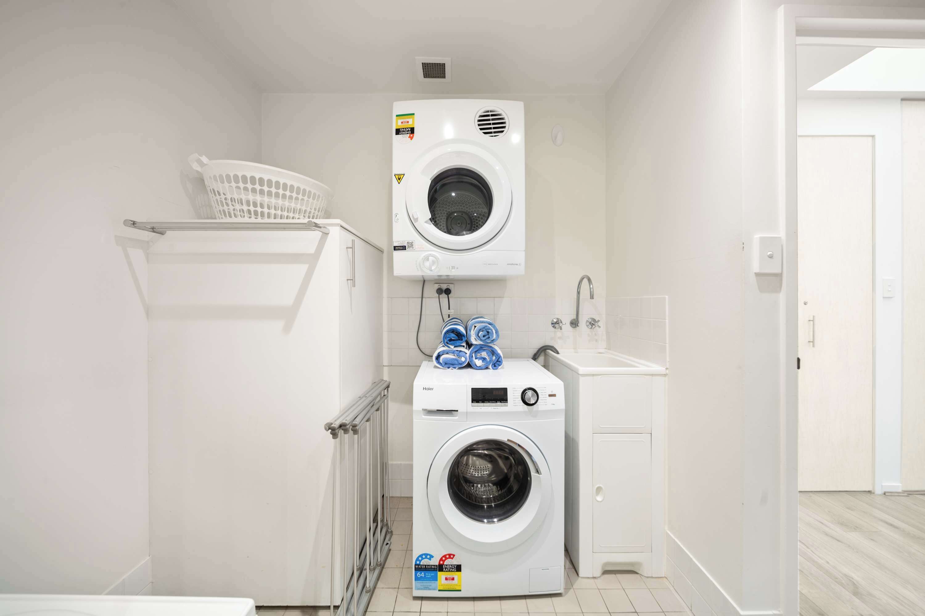 Une petite buanderie équipée d’un lave-linge et d’un sèche-linge à chargement par le haut, d’un petit évier et d’un panier posé sur l’étagère.