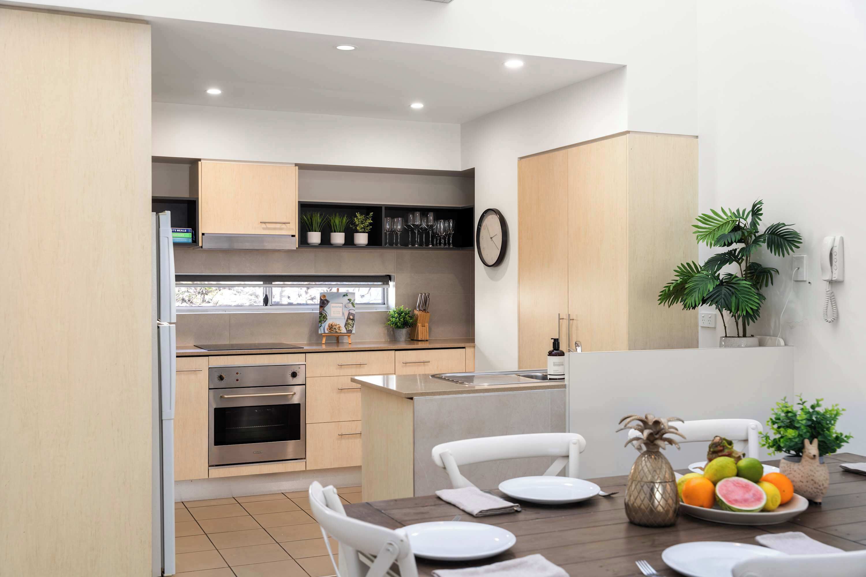 A modern kitchen with built-in appliances, a dining area with white chairs, and a fruit bowl on the table.