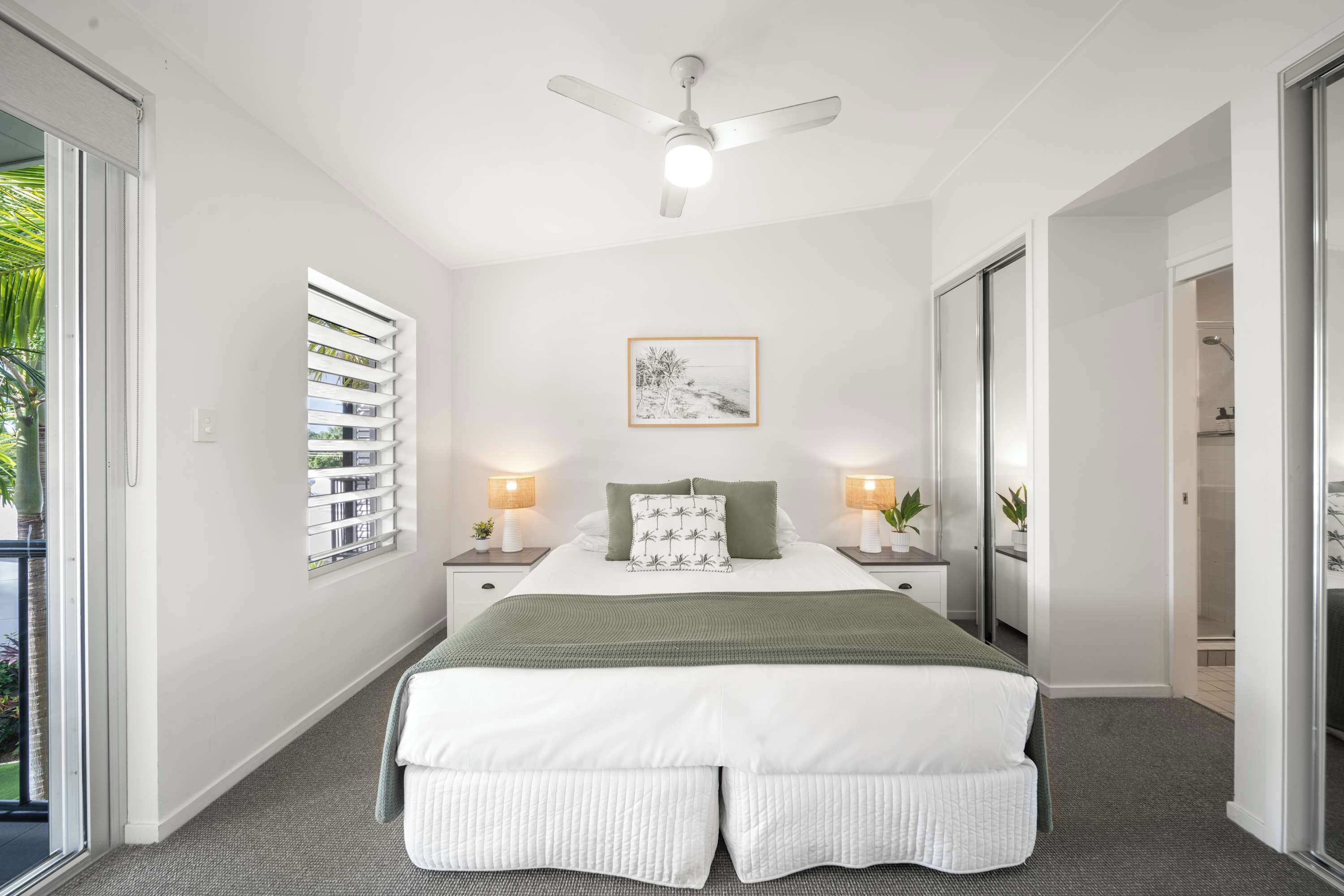 A bedroom with a bed, a ceiling fan, a window with blinds, bedside lamps, and a framed picture on the wall.
