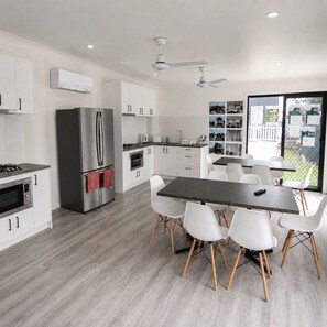 Eine moderne Küche mit weißen Schränken, einem Edelstahl-Kühlschrank, einem Esstisch mit Stühlen und Blick auf einen Garten durch große Fenster.