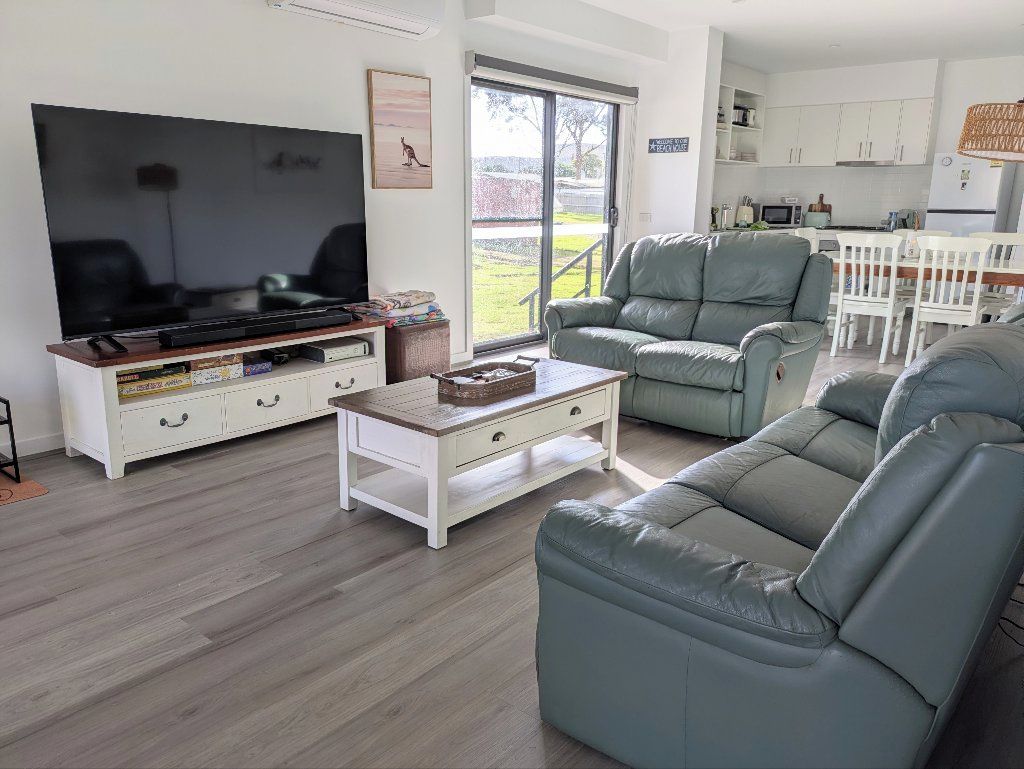 Ein Wohnzimmer mit Flachbildfernseher, einer Couch, einem Couchtisch und einem Essbereich mit weißem Tisch und Stühlen.