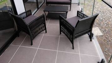 A balcony with a seating area consisting of two sofas and two chairs, a glass railing, and a tiled floor.