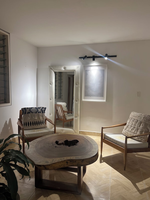 A room with a round wooden table, two upholstered chairs with patterned cushions, a doorway leading to another room, and a wall-mounted light fixture.