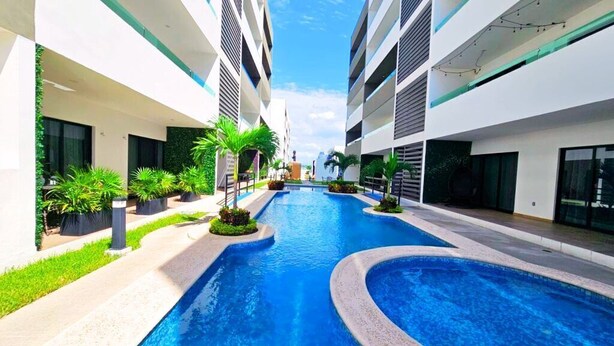 A modern hotel with a central swimming pool flanked by multi-story buildings, surrounded by greenery and potted plants. - Marina Cerritos 507 Coastal Retreat – 2BR (Mazatlan)