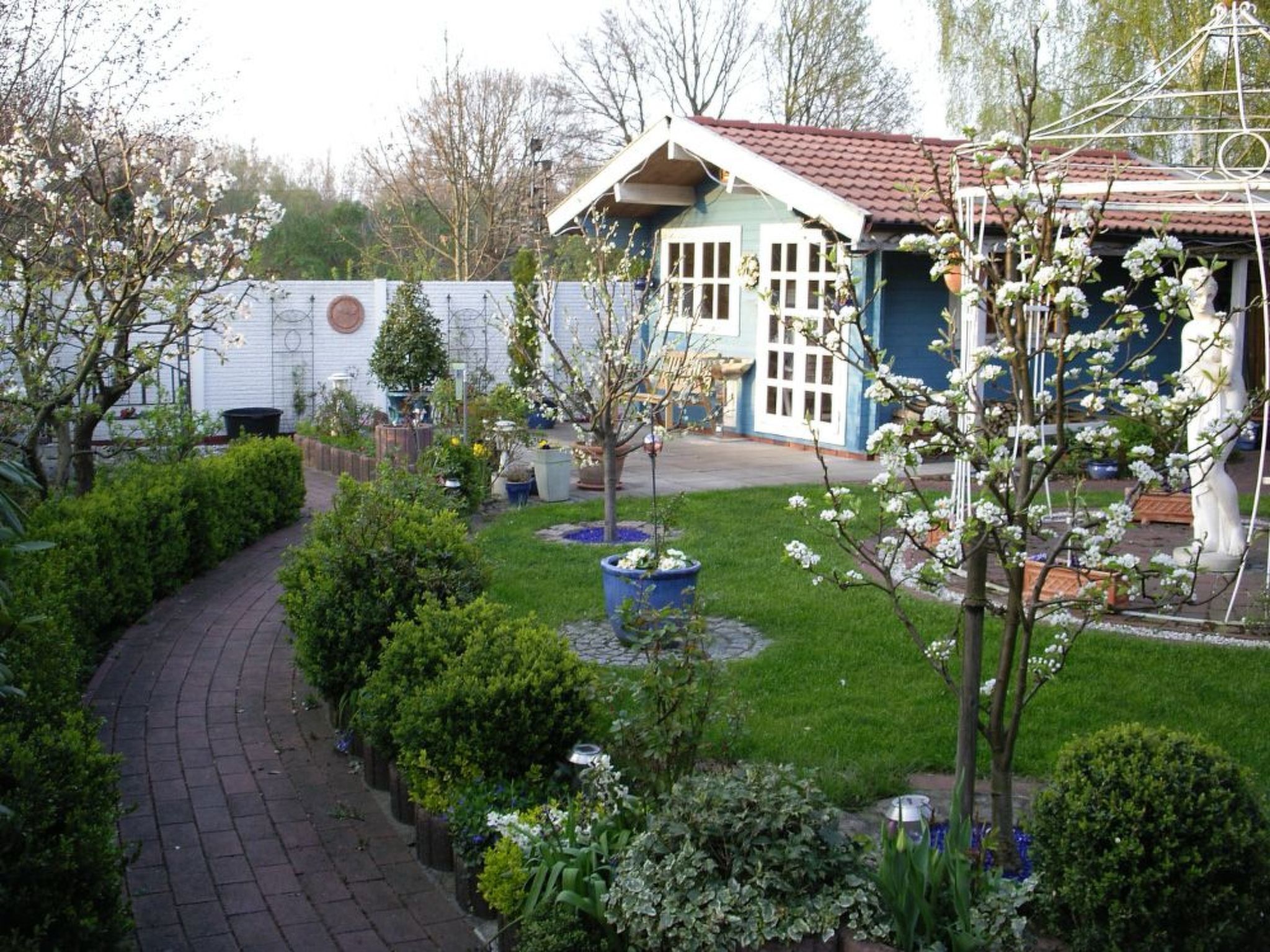 Ein Garten mit gepflastertem Weg, unterschiedlicher Bepflanzung und einem Gebäude mit rotem Dach.