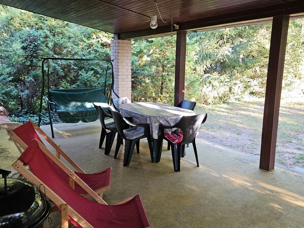 An outdoor covered patio with a table and chairs, surrounded by greenery.