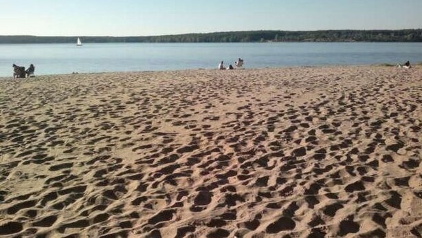 En sandstrand med fotspår, ett stilla vatten och en segelbåt i fjärran.