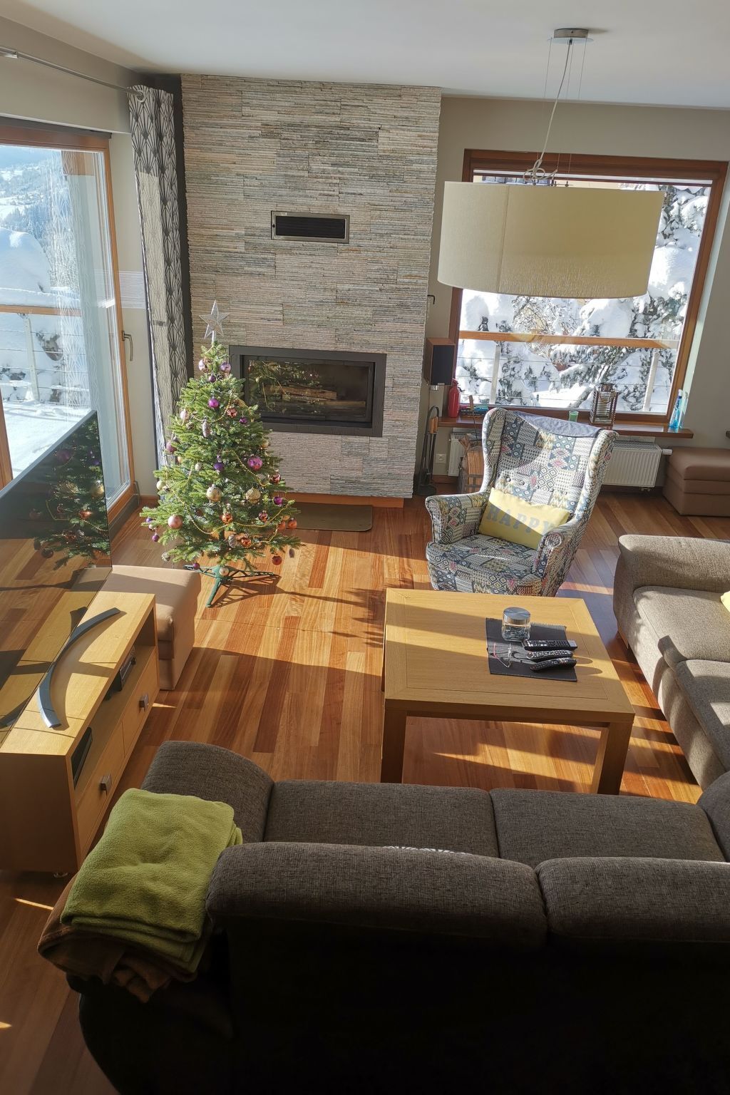 Un salon doté d’une cheminée en pierre, d’un sapin de Noël et d’une grande fenêtre offrant une vue sur des arbres couverts de neige.