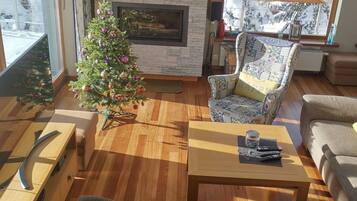 Un salon doté d’une cheminée en pierre, d’un sapin de Noël et d’une grande fenêtre offrant une vue sur des arbres couverts de neige.