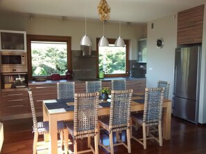 A modern kitchen with wooden cabinetry, a built-in oven, a stainless steel refrigerator, and a dining area with wicker chairs and a wooden table.
