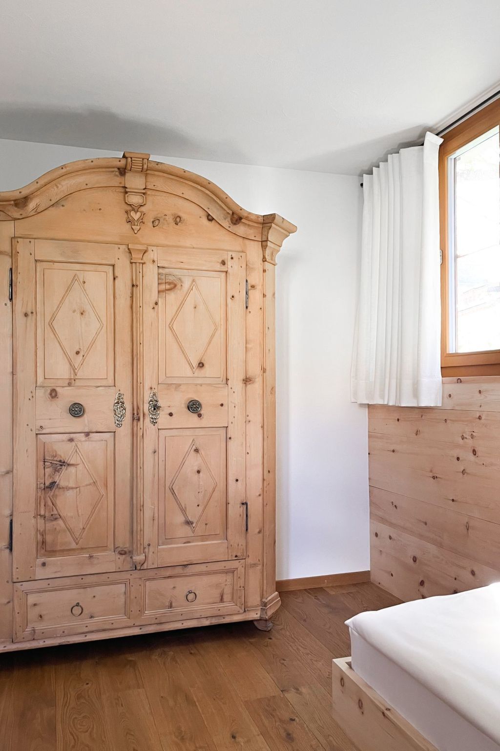 Un armario de madera con dos puertas y un cajón pequeño, una cama de madera con colchón blanco, y una ventana con cortinas blancas.