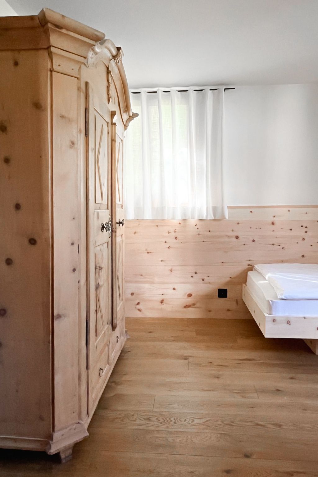 A room with a wooden wardrobe, a bed, and a window with sheer curtains.