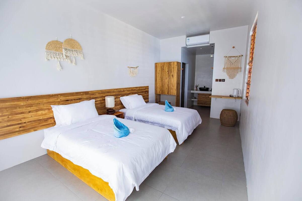 Habitación de hotel con dos camas, cabecero de madera, mesita de noche pequeña con lámpara y unidad de aire acondicionado montada en la pared.