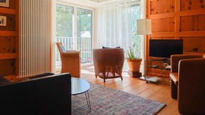 Ein Wohnzimmer mit Fernseher, Rattanmöbeln, einem Topfpflanzen und Blick ins Freie.