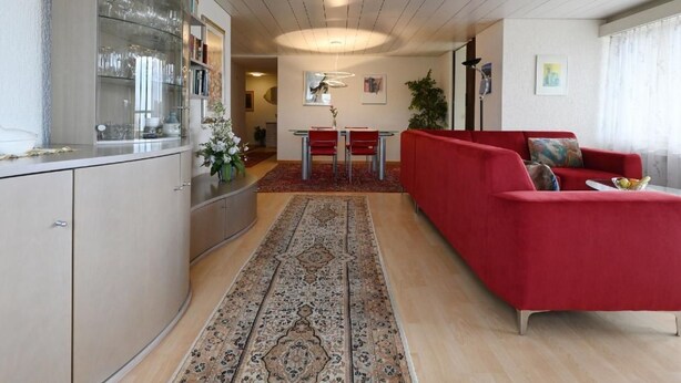 Een moderne woonkamer met een rode bank, een eethoek met een rode tafel en stoelen, en een keuken met een glazen kast.