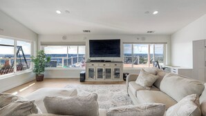 Ein geräumiges Wohnzimmer mit einem großen Flachbildfernseher, einer gemütlichen Sofagarnitur und Blick auf die Stadt durch große Fenster.
