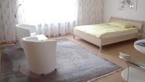 A modern hotel room with a bed, a white armchair, a small table, and a large window with sheer curtains. - Appartement Alpenblick / G6 by Interhome (Weggis)