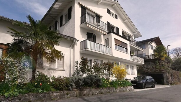 A multi-story residential building with balconies, a car parked in front, and a stone wall with greenery. - Haus Stirnemann/ Wohnung M-2 by Interhome (Weggis)