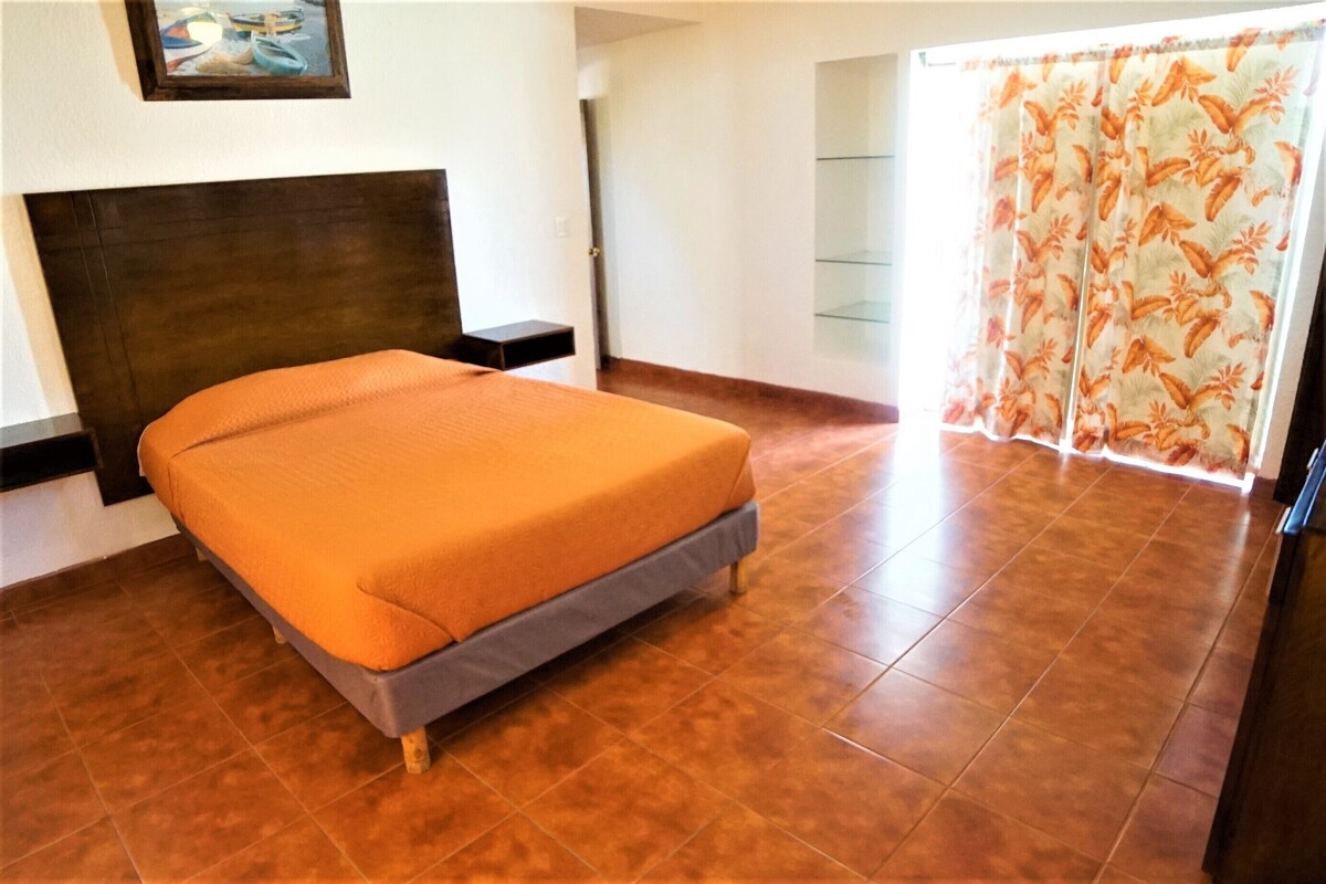 Um quarto com cama, cabeceira, janela com cortinas e porta.