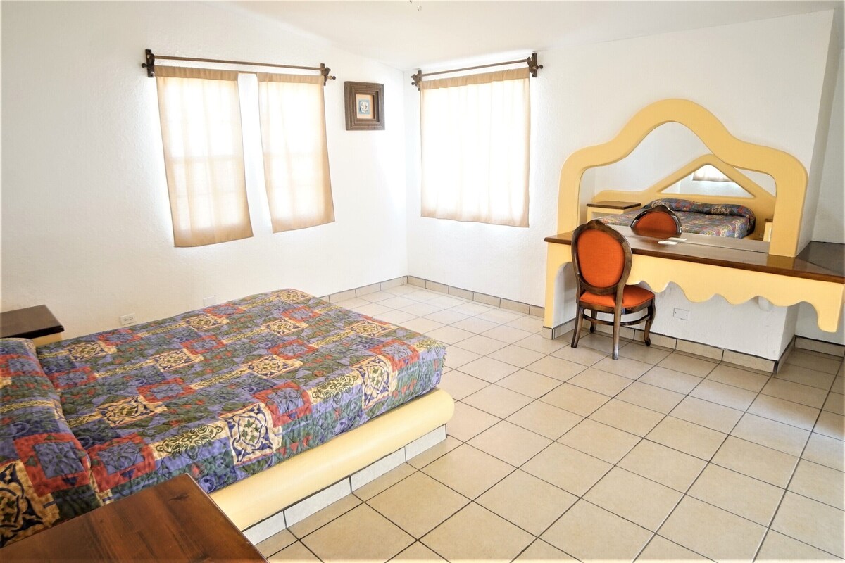 Quarto com cama, escrivaninha e duas janelas com cortinas.