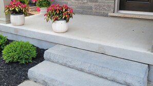 Una casa con facciata in pietra, una porta d'ingresso e un piccolo portico con gradini che conducono all'entrata. Sul portico ci sono tre vasi con piante a fiori rosa.