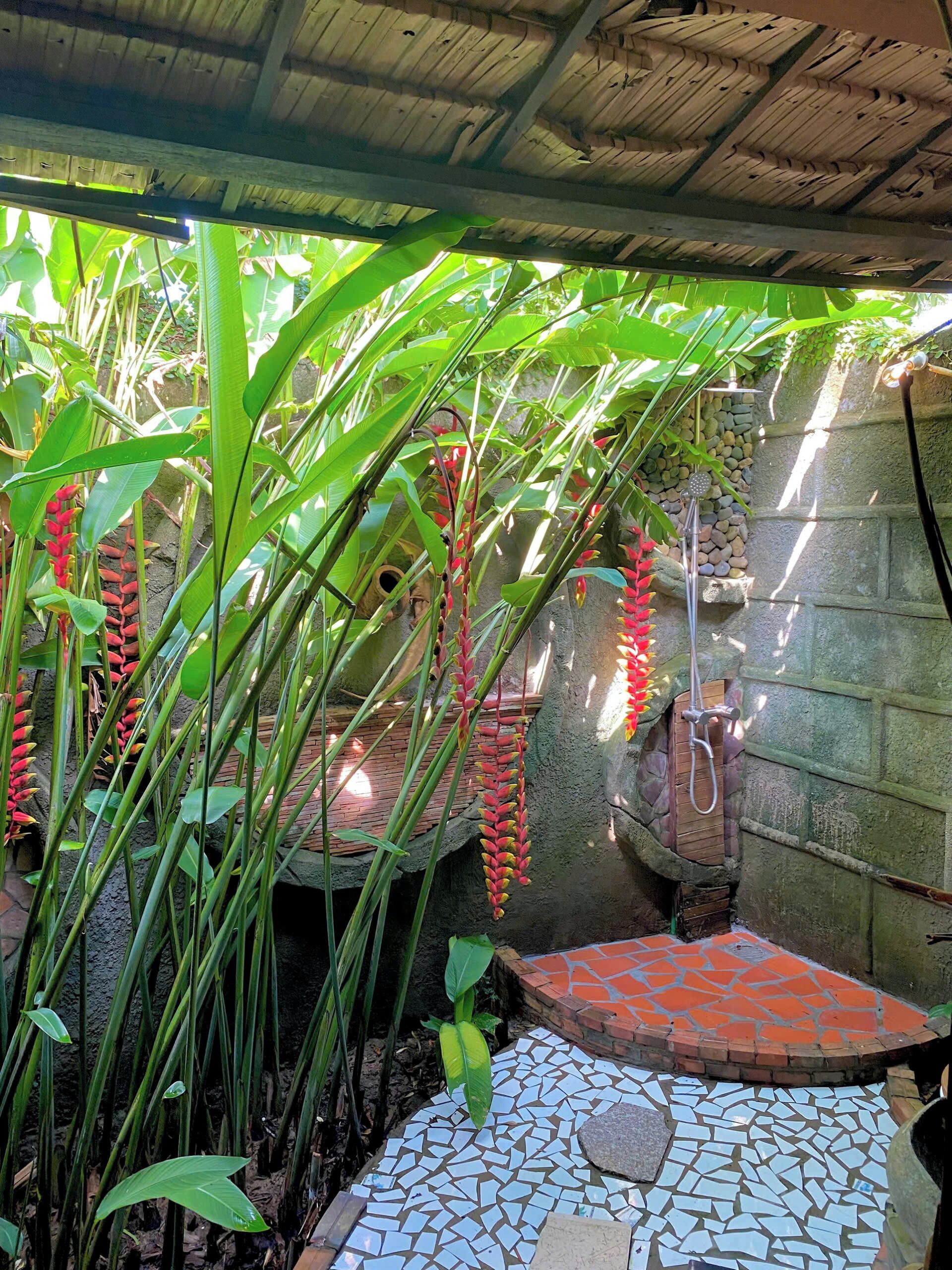A covered outdoor area with a tiled floor, surrounded by lush greenery and a stone wall.
