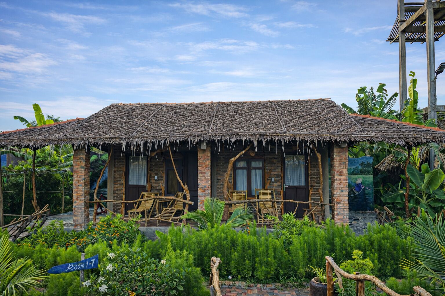 Een rustiek hutachtig gebouw met rieten dak, bakstenen muren en een veranda met houten stoelen.