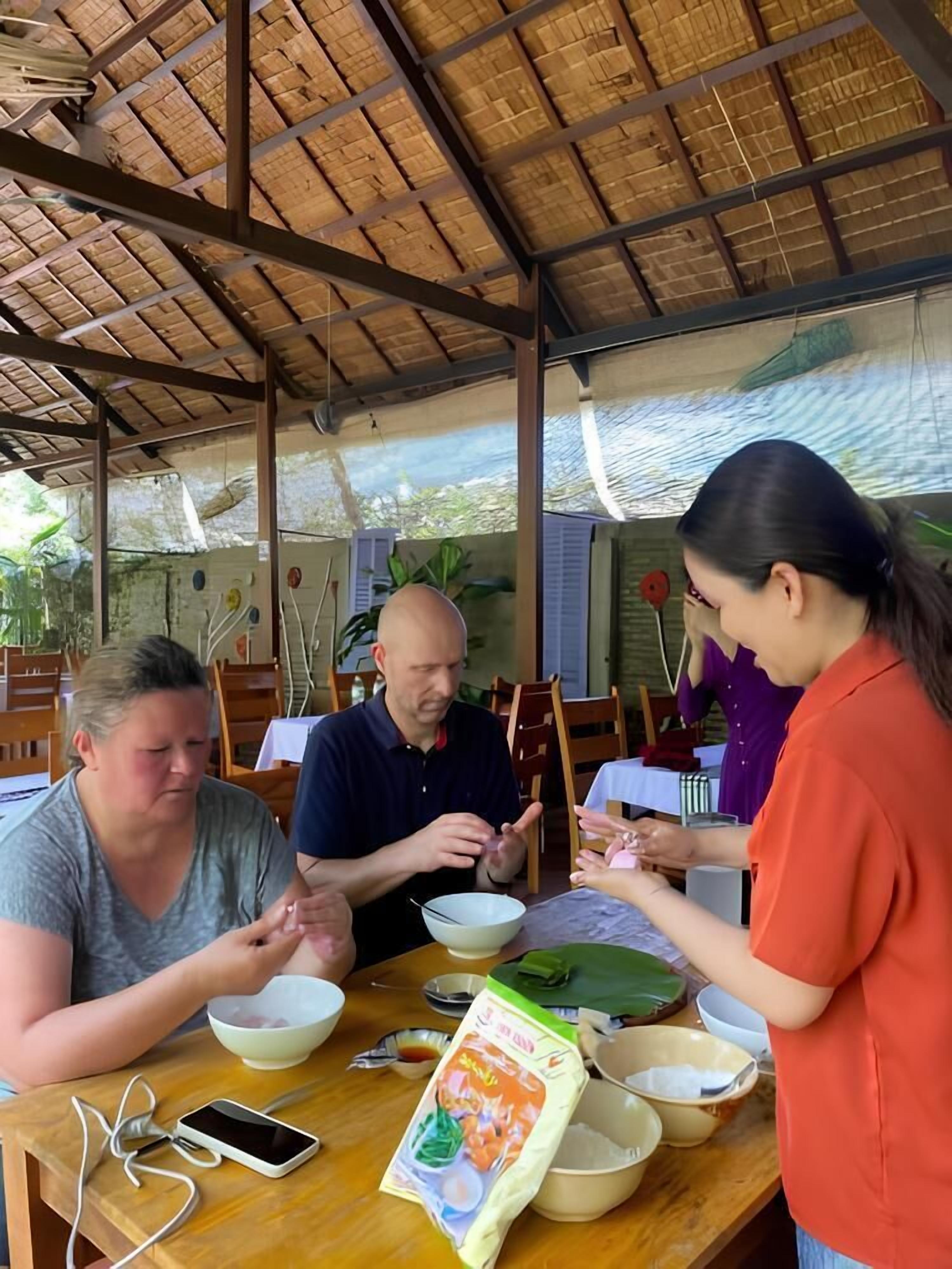 Uma área de refeições com telhado de palha, mesas e cadeiras de madeira. Há tigelas, um smartphone e um pacote de comida na mesa.
