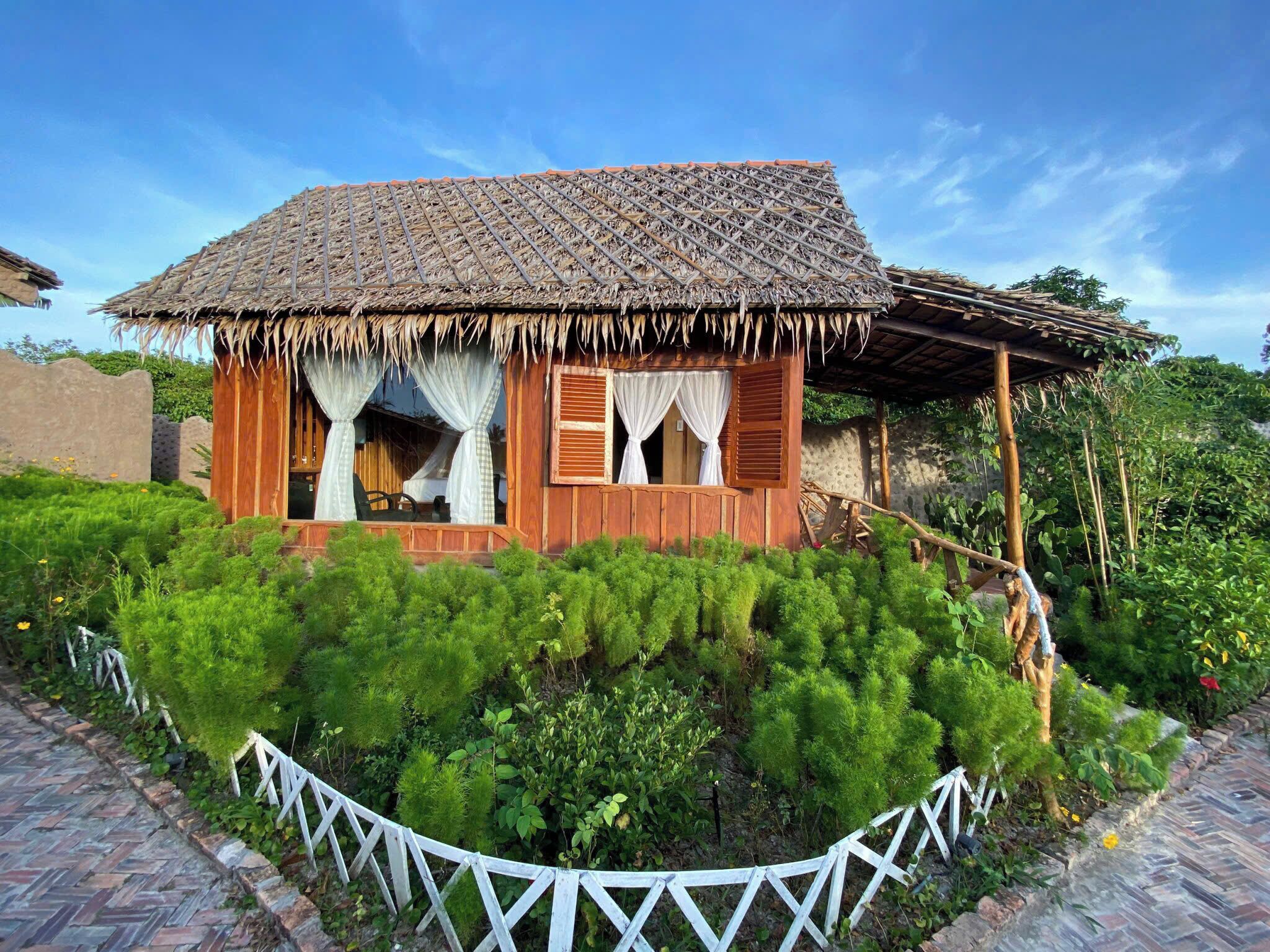 Une maison rustique en bois, avec un toit de chaume, entourée d’une végétation luxuriante et d’une clôture blanche en bois.