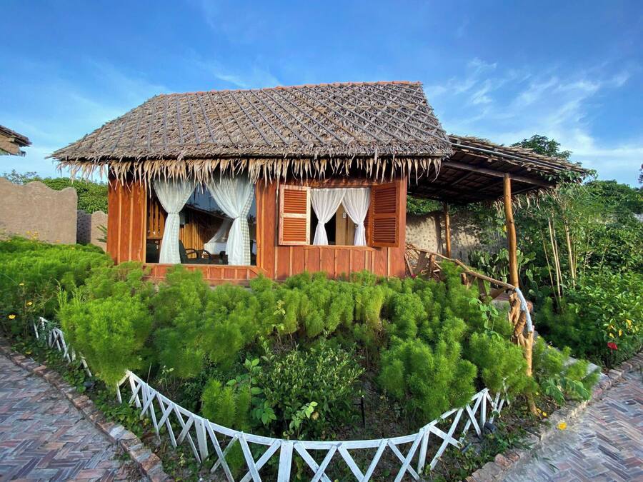 Une maison rustique en bois, avec un toit de chaume, entourée d’une végétation luxuriante et d’une clôture blanche en bois.