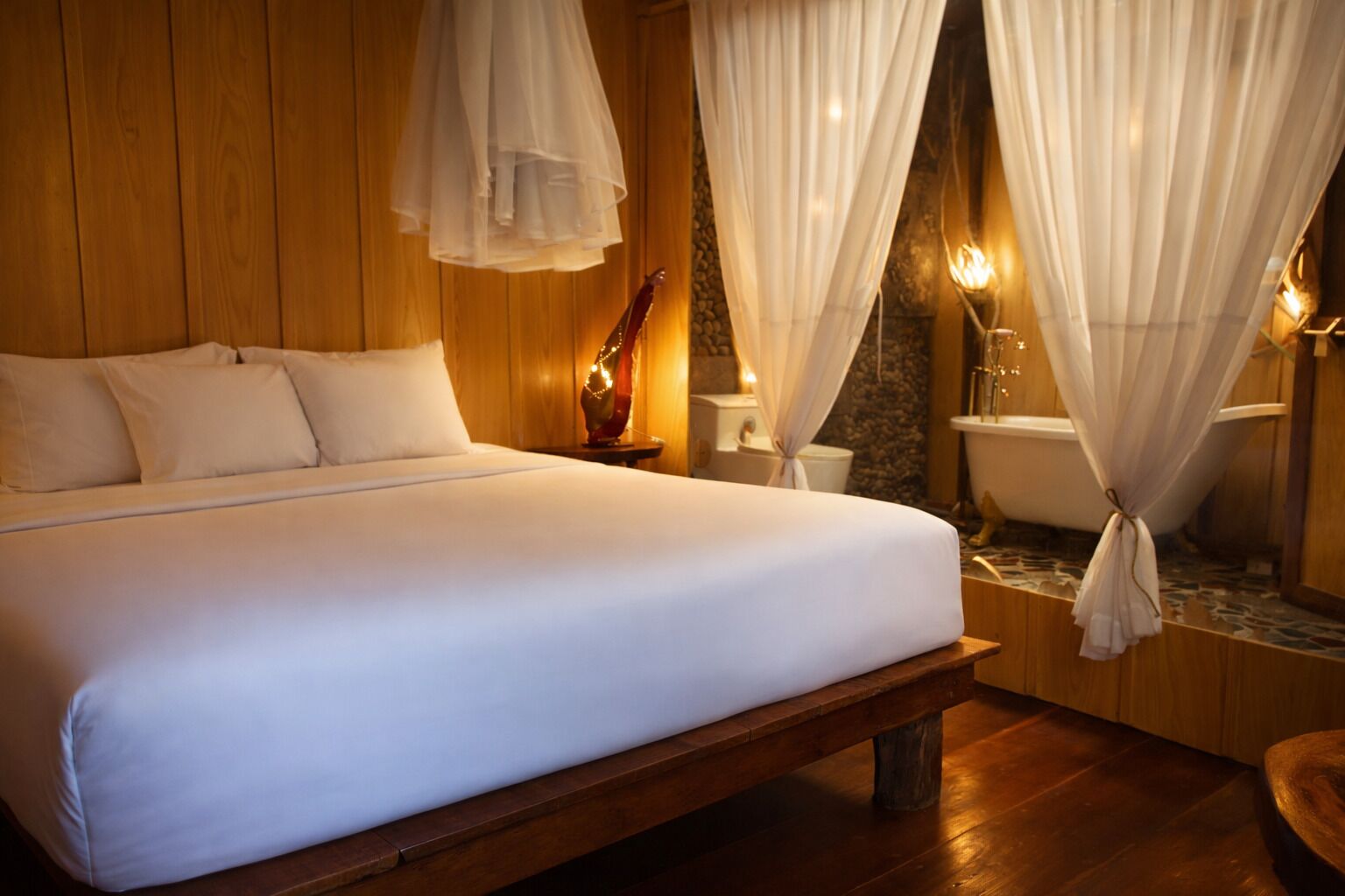 Een bed met witte lakens, een houten hoofdeinde en een bank aan het voeteneinde. Er is een nachtkastje met een lamp en een spiegel. De kamer heeft houten wanden en een accentmuur van steen.