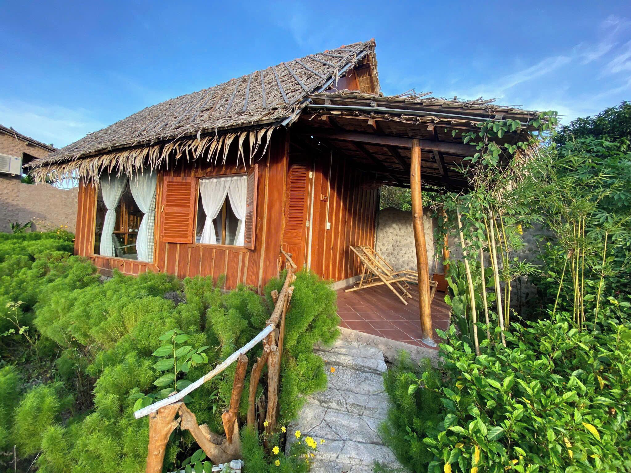 A rustic wooden house with a thatched roof, surrounded by lush greenery and a stone pathway.