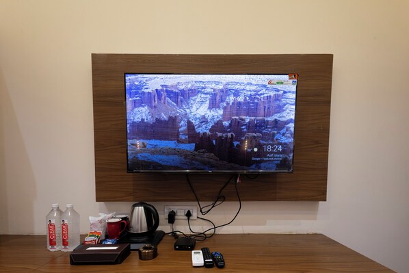 A television mounted on a wall displaying a snowy landscape, a wooden table with a coffee maker, two water bottles, a tissue box, and a remote control. - Lotus Feet Inn (Varanasi)