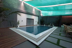 An indoor swimming pool with a brick wall, a wooden deck, and a covered area with chairs. - Lotus Feet Inn (Varanasi)