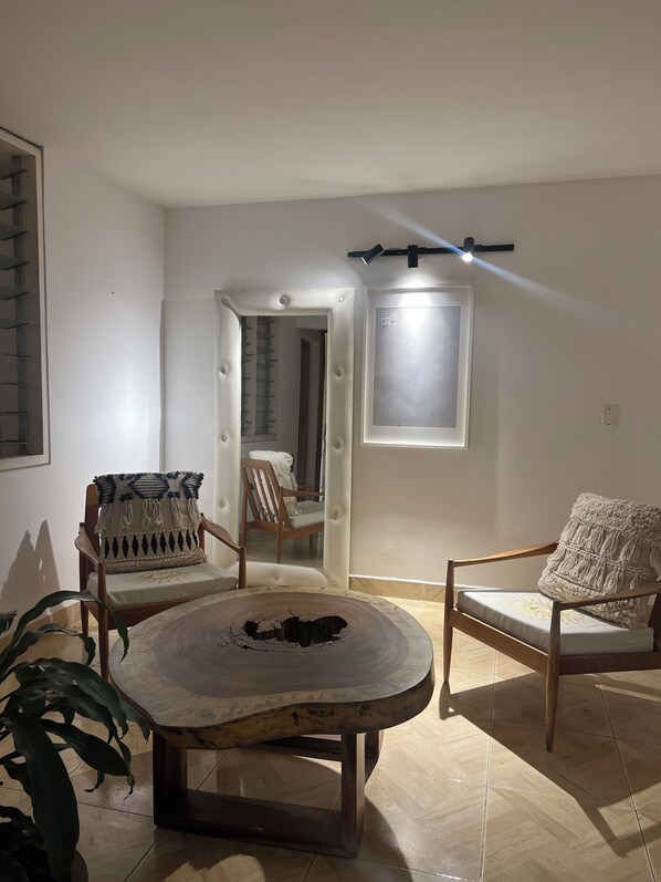 Une pièce comprenant une table ronde en bois, deux fauteuils rembourrés avec des coussins à motifs, une porte-fenêtre donnant sur une autre pièce et un luminaire fixé au mur.