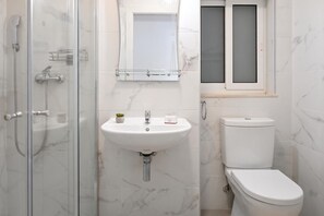 A bathroom with a glass-enclosed shower, a white sink with a marble backsplash, a toilet, and a window with frosted glass.