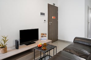 A modern room with a flat-screen TV, a wooden coffee table with fruits and a glass, a leather sofa, and a closed door with a 'No warning' sign.