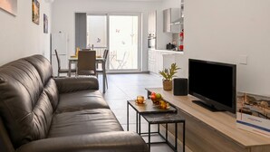 A modern living room with a leather sofa, a flat-screen TV, a small coffee table, and a dining area with a table and chairs. - Camelia Seaview 1 Bd Apart In St Paul Bay (St. Paul's Bay)