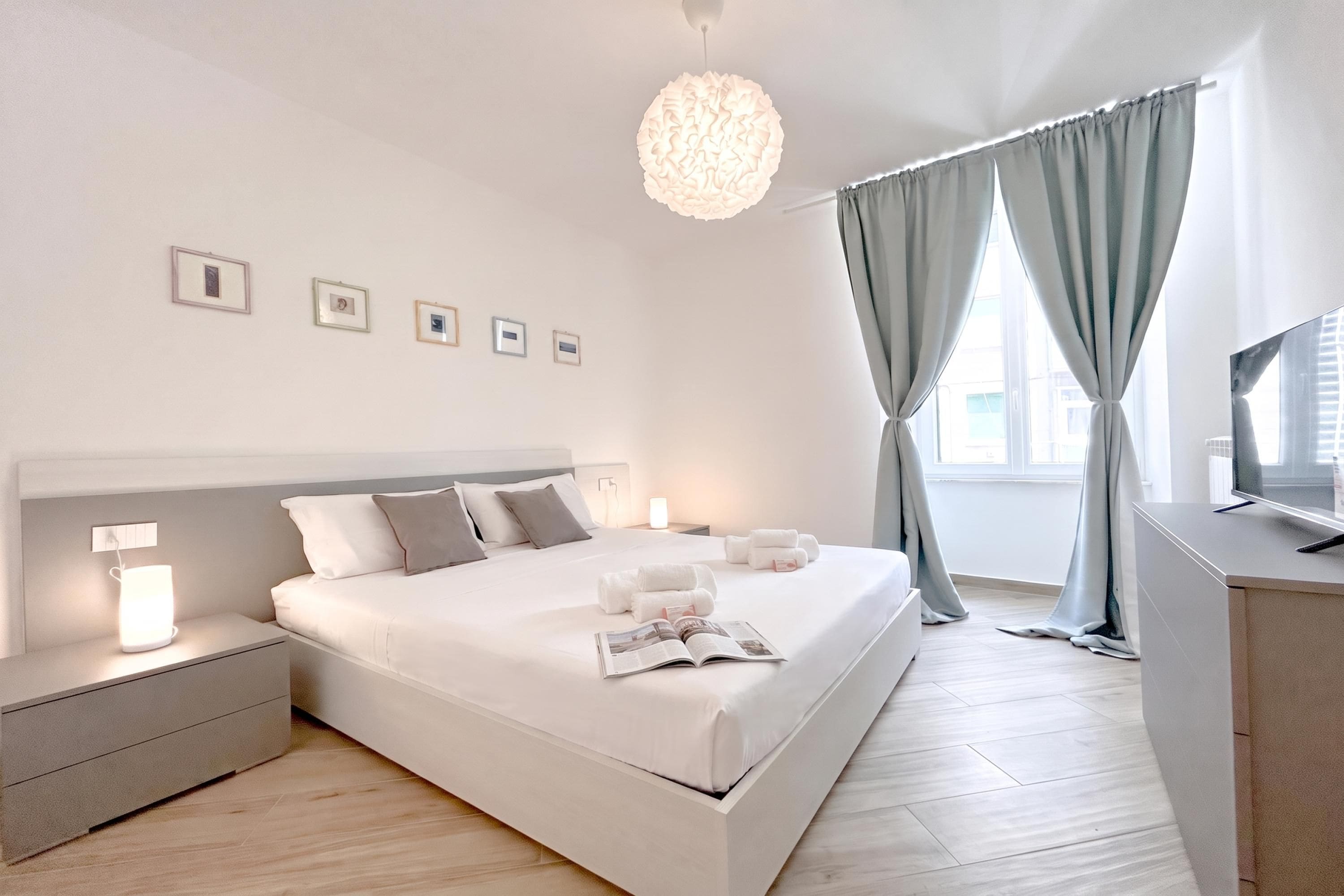 A modern bedroom with a large bed, bedside tables, a reading lamp, a flat-screen TV, and a window with curtains.