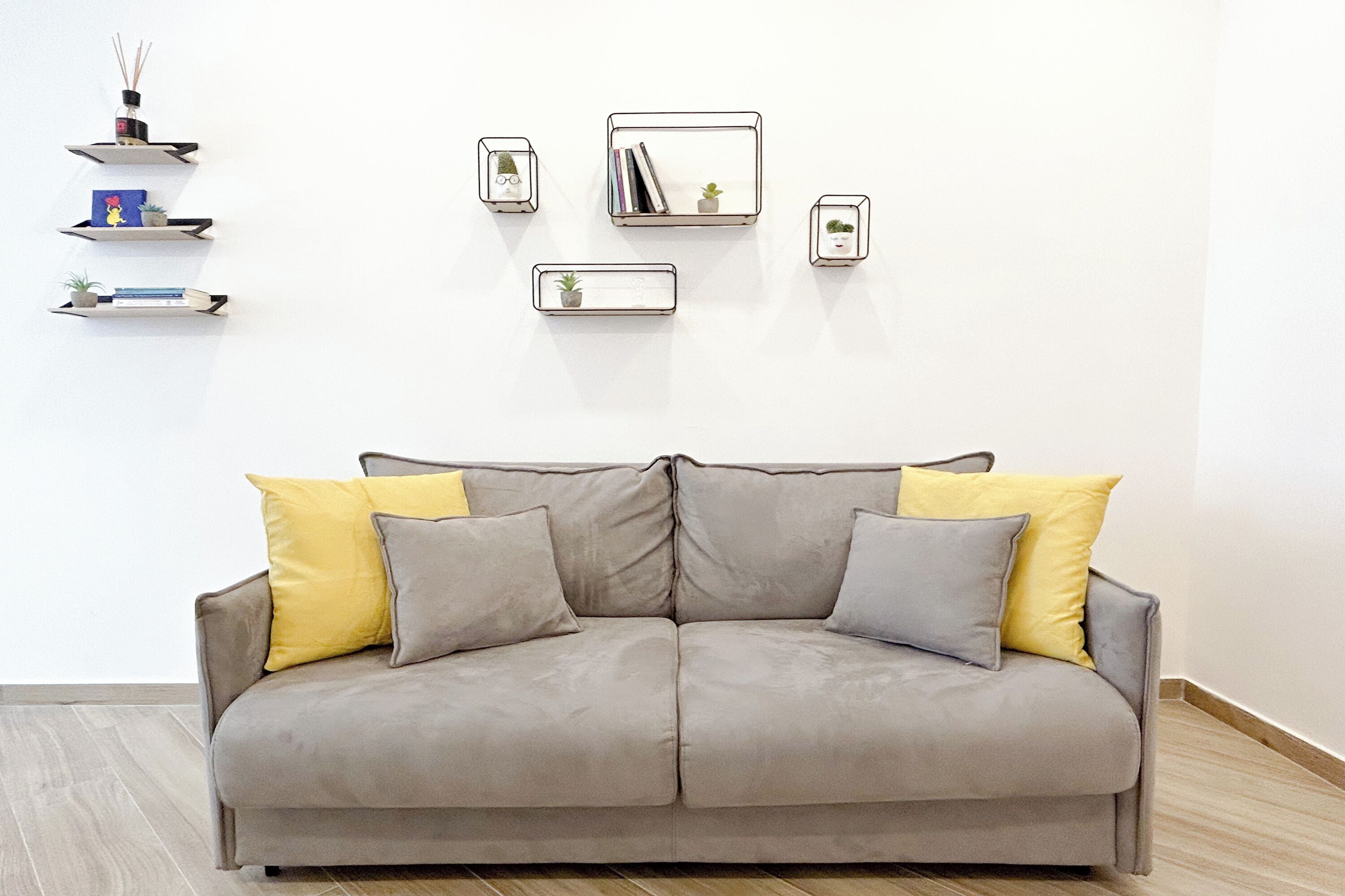 A grey sofa with two yellow cushions, a white wall, and three shelves with various items.