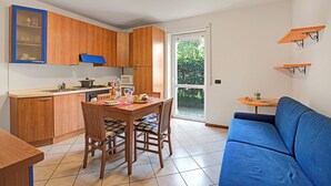A compact kitchen with wooden cabinets, a dining table with chairs, a blue sofa, and a small table with a cup. - Appartamento Gioale (Riva del Garda)