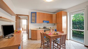 A modern kitchen with wooden cabinets, a dining table with chairs, a TV, and a large window. - Appartamento Gioale (Riva del Garda)