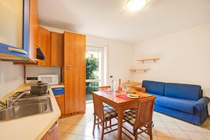 A compact kitchen with a dining area, a blue sofa, and a view through the door. - Appartamento Gioale (Riva del Garda)