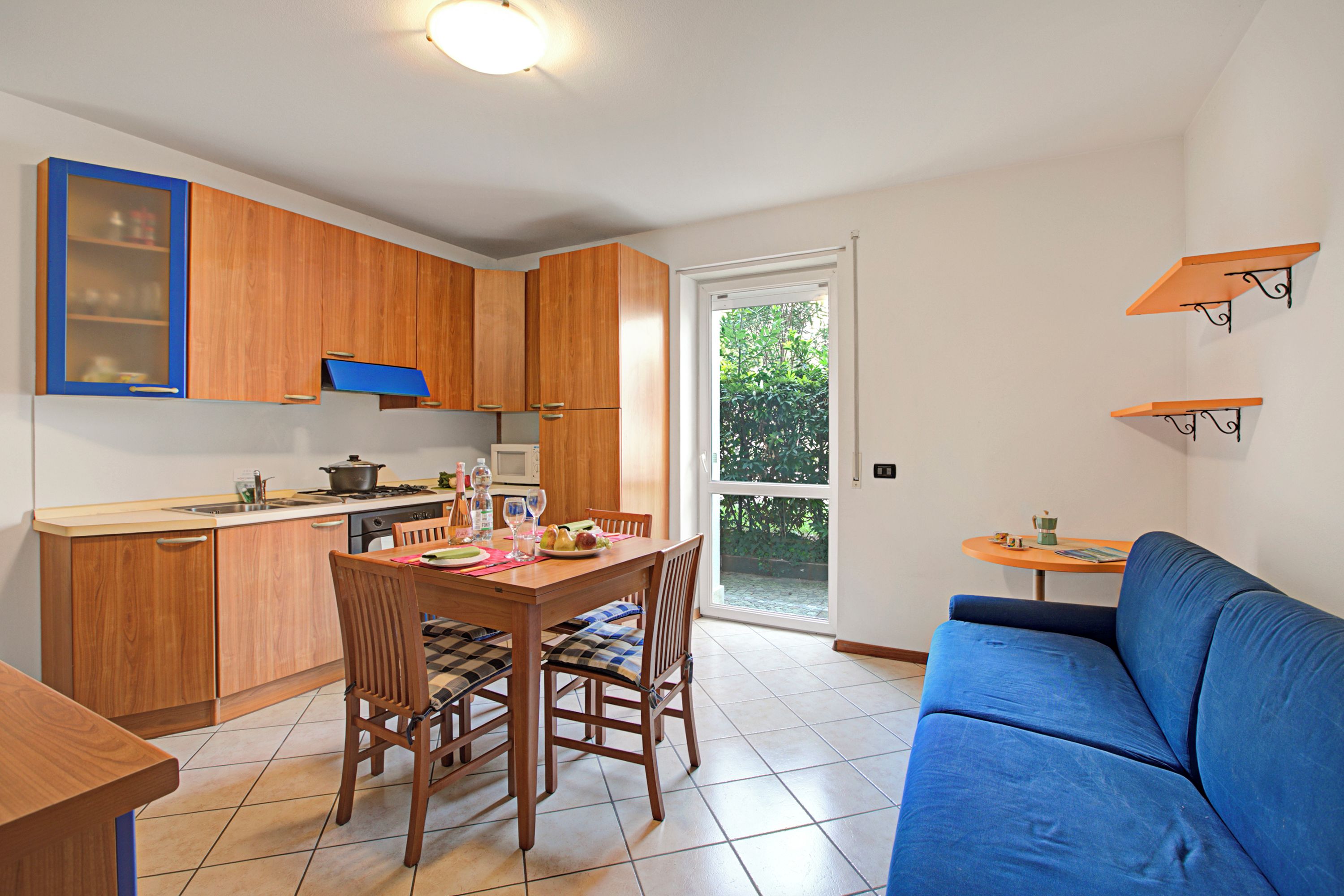 A compact kitchen with wooden cabinets, a dining table with chairs, a blue sofa, and a small table with a cup.