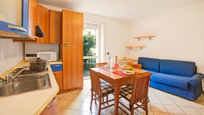 A compact kitchen with a dining area, a blue sofa, and a view through the door. - Appartamento Gioale (Riva del Garda)