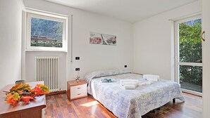 A bedroom with a bed, bedside table, radiator, window, and a view of greenery. - Appartamento Gioale (Riva del Garda)