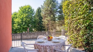 Una mesa redonda con mantel estampado, rodeada de sillas blancas, ubicada al aire libre con vista a árboles y una cerca.