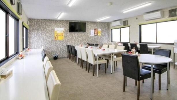 Uma sala de conferências com várias mesas e cadeiras, uma TV de tela plana na parede e grandes janelas que proporcionam luz natural.