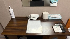 Quarto de hotel com escrivaninha de madeira, cadeira de escritório preta, controle remoto, caixa de lenços, frasco de álcool em gel e saco plástico.